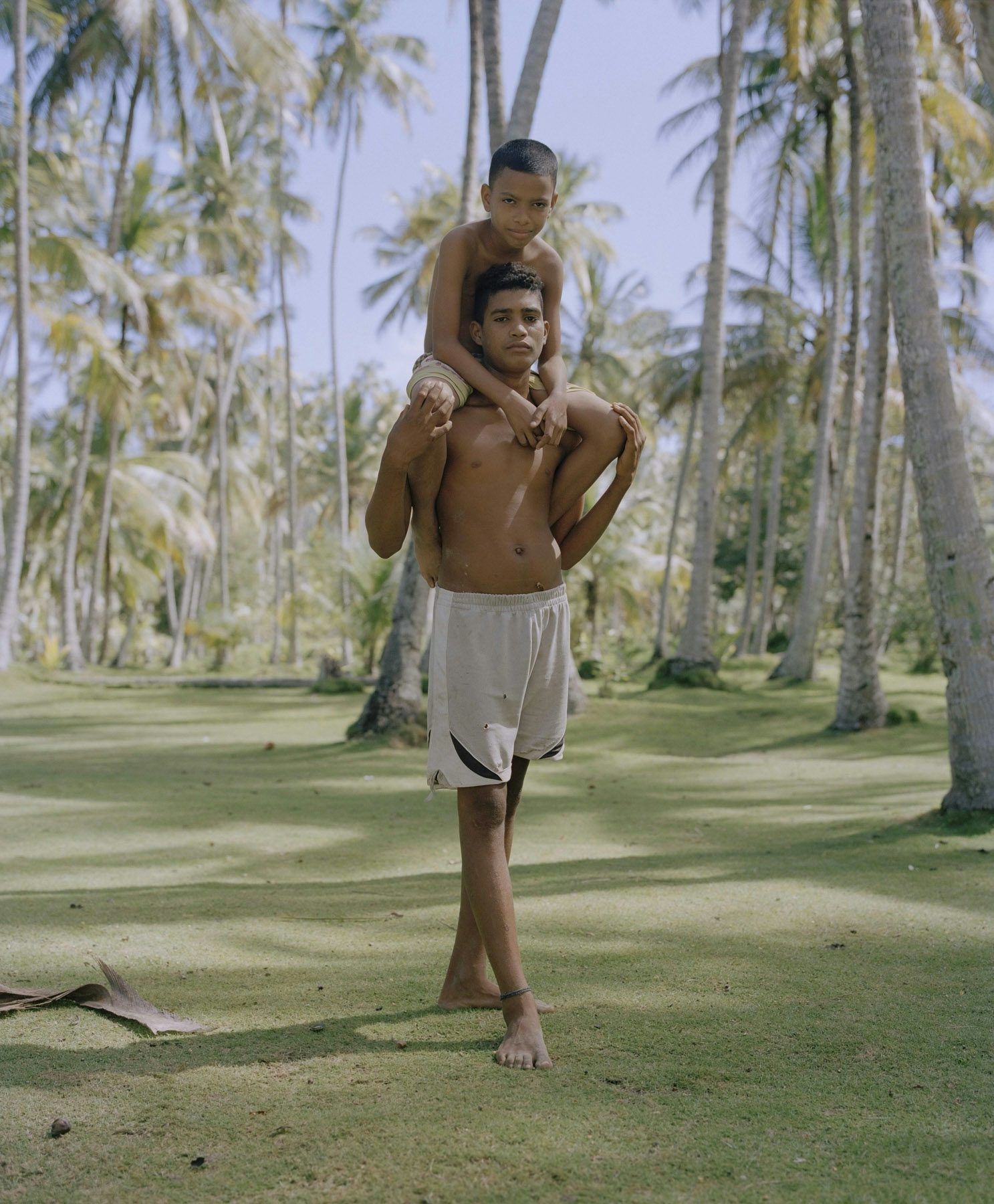 Image by Silvana Trevale of a young shirtless man stood among trees with a child on his shoulders
