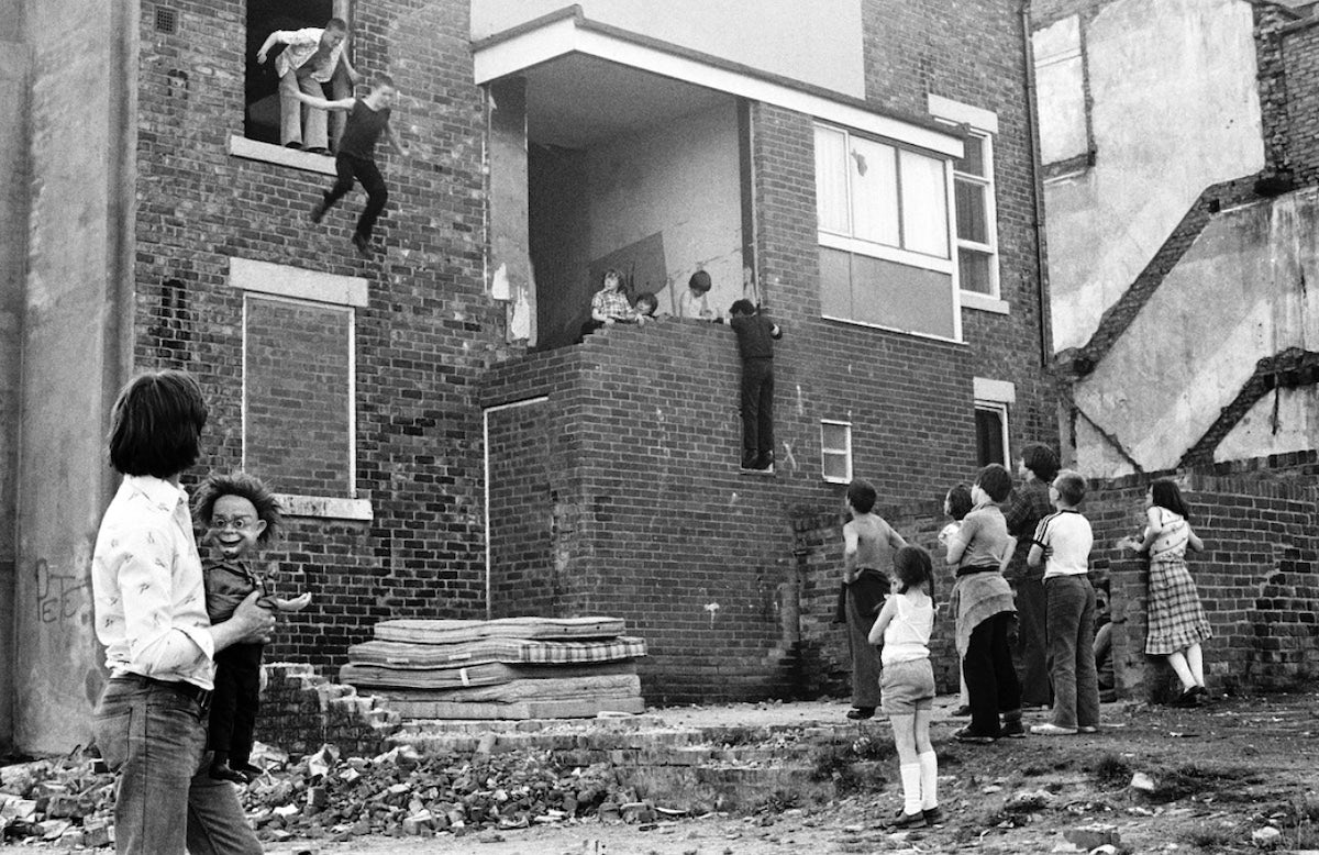 Black and white photograph by Tish Murtha showing a child jumping from a window onto a pile of mattresses