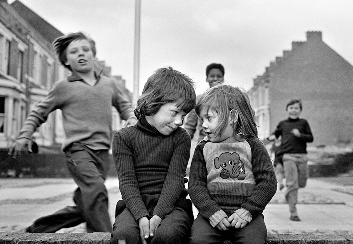 Black and white photograph by Tish Murtha showing two children sat on a wall smiling at one another
