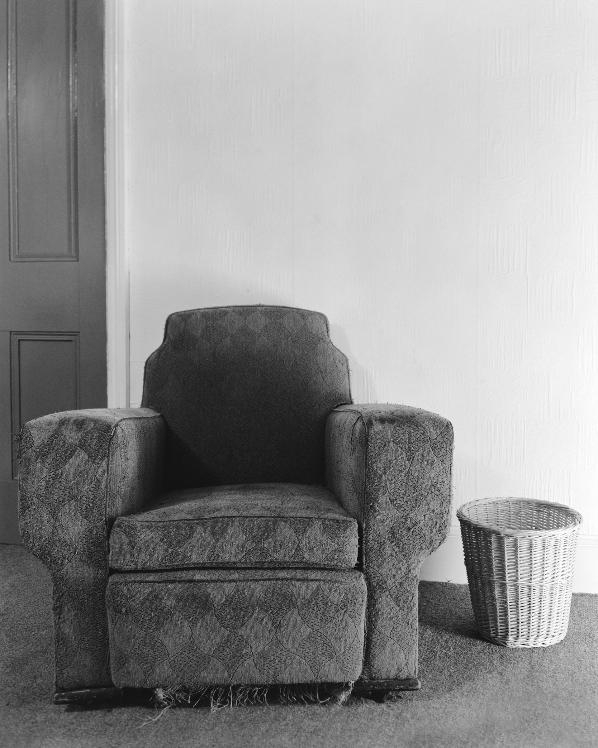 Black and white photograph of an empty armchair next to a wastepaper bin, which features in Life As It Is by John Myers
