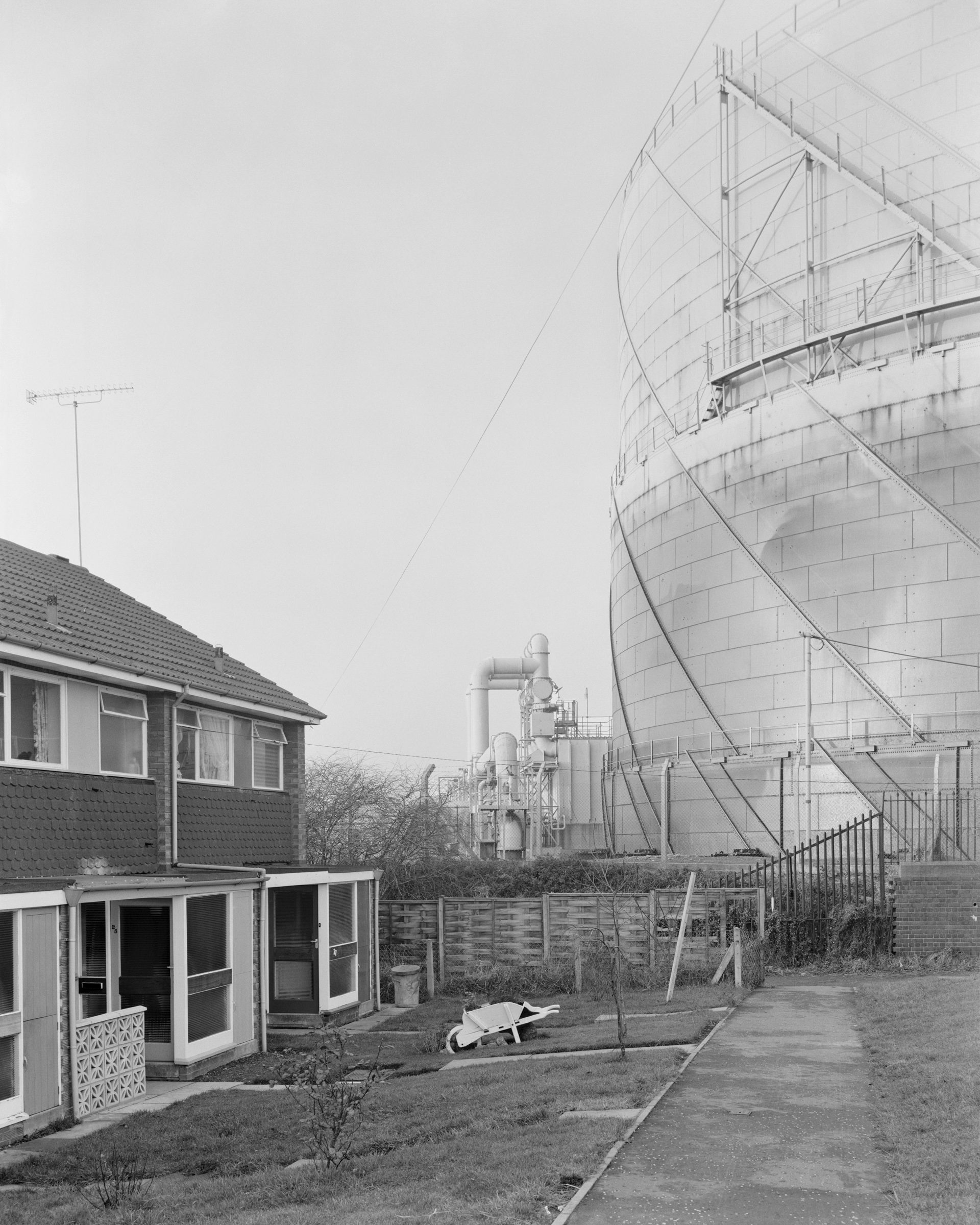 Black and white photograph of a house next to a gasometer, which features in Life As It Is by John Myers