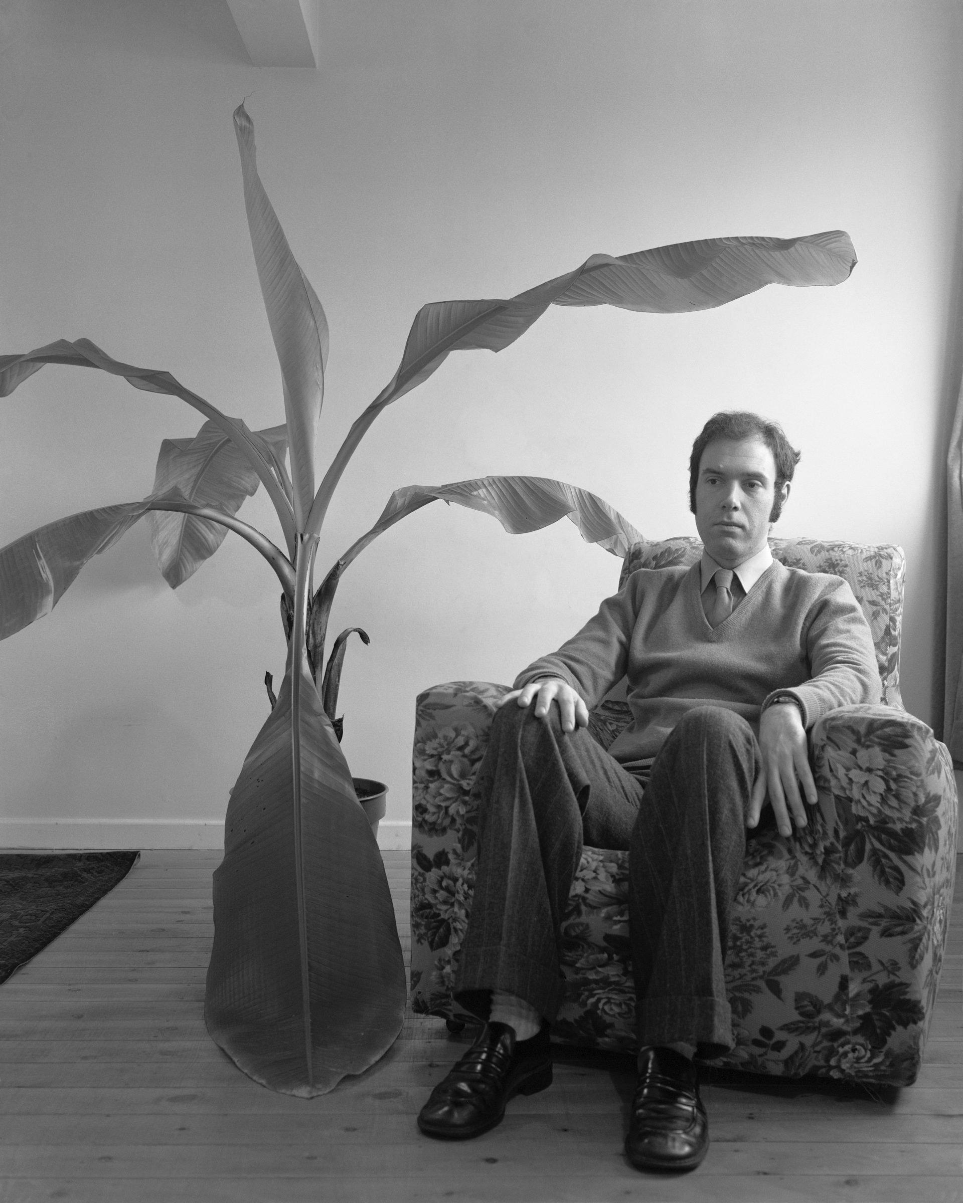 Black and white photograph of a man sat in an armchair next to a banana plant, which features in Life As It Is by John Myers