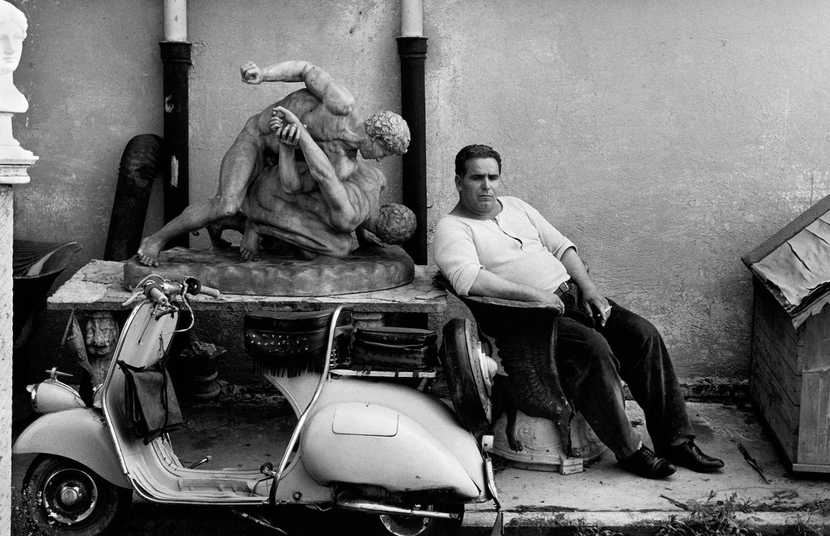 Black and white photograph of a man sat on a chair in Rome next to a statue, as part of the new William Klein exhibition called YES at the ICP