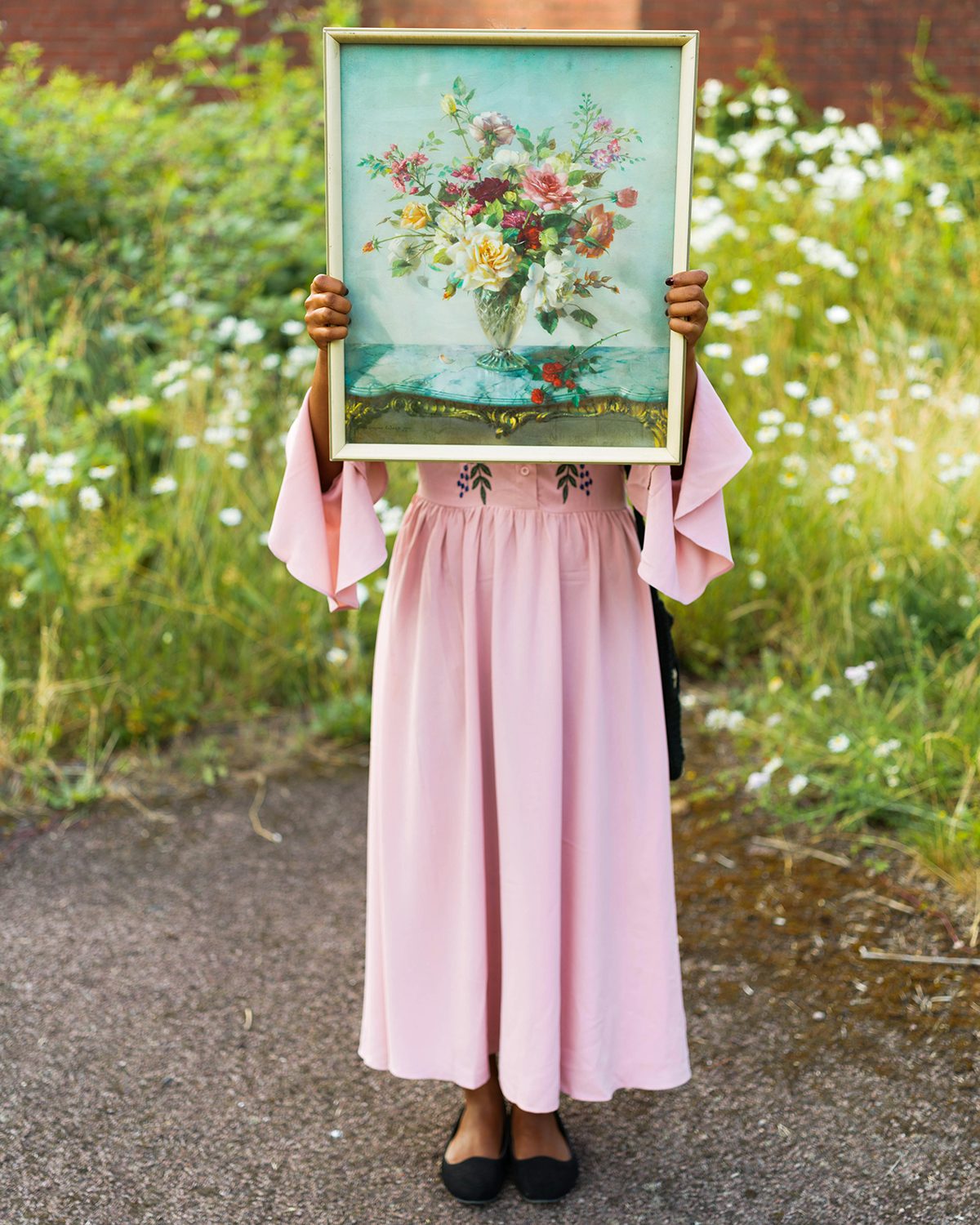 Image shows a person in a pink outfit holding a picture of a vase of flowers over their face, taken from Kavi Pujara's book This Golden Mile