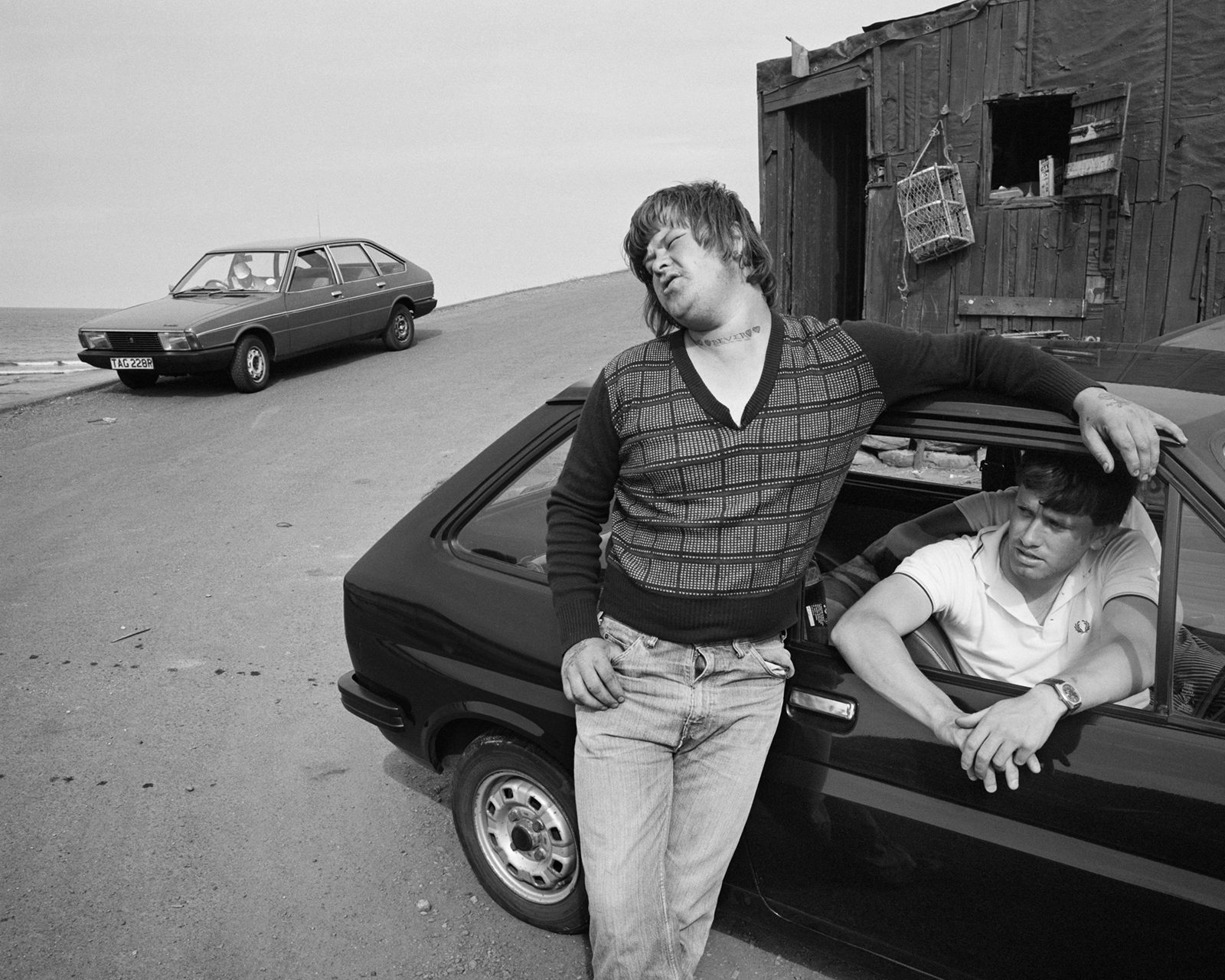 Black and white photograph of two people hanging out by a car, which appears in the new Chris Killip exhibition