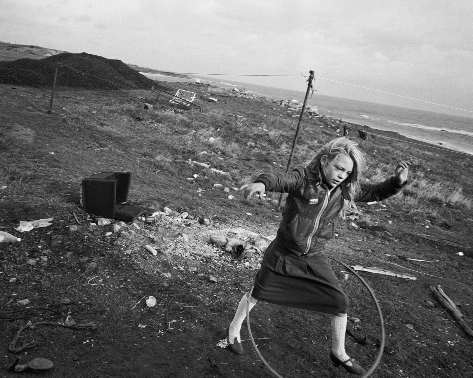 Black and white photograph of a young person playing with a hula hoop next to the coast, which appears in the new Chris Killip exhibition