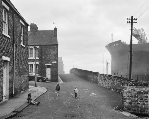 Black and white photograph of three children in the street with a ship docked next to it, which appears in the new Chris Killip exhibition