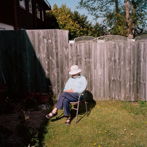 Image shows Pauline sitting on a chair in her garden, wearing a hat concealing her face, photographed by Taylor Wessing prize winner 2022, Clementine Schneidermann