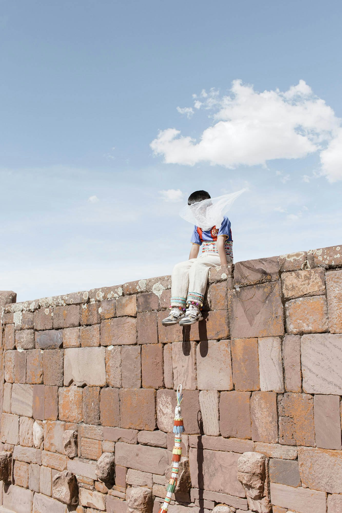 Image by River Claure showing a young boy with his face covered my material, sat atop a wall