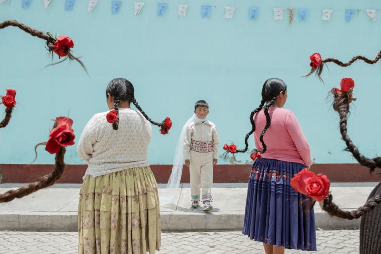 Image by River Claure showing a young boy stood on a pavement against a blue wall, surrounded by people whose braided hair juts into the frame