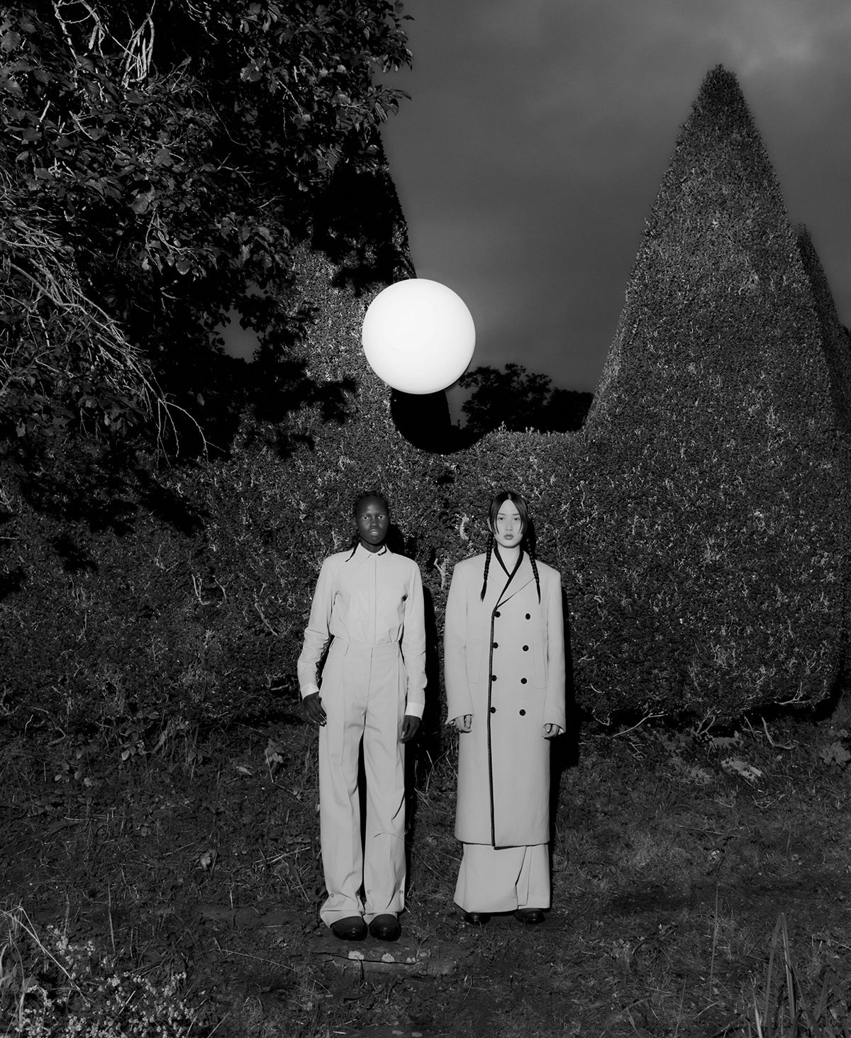 Black and white image by Agnes Lloyd-Platt shows two people in light-coloured suits stood side by side against a hedgerow, with a moon-shaped ball floating overhead