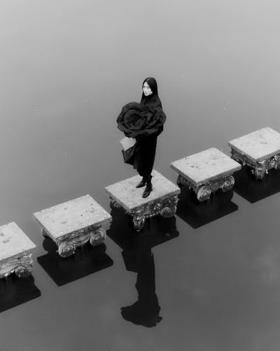 Black and white image by Agnes Lloyd-Platt shows a person wearing a garment in the shape of a flower head, stood in the middle of a row of square steps leading over water