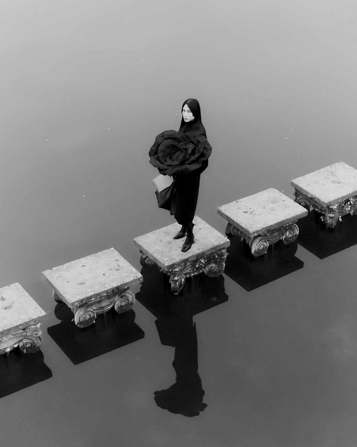 Black and white image by Agnes Lloyd-Platt shows a person wearing a garment in the shape of a flower head, stood in the middle of a row of square steps leading over water