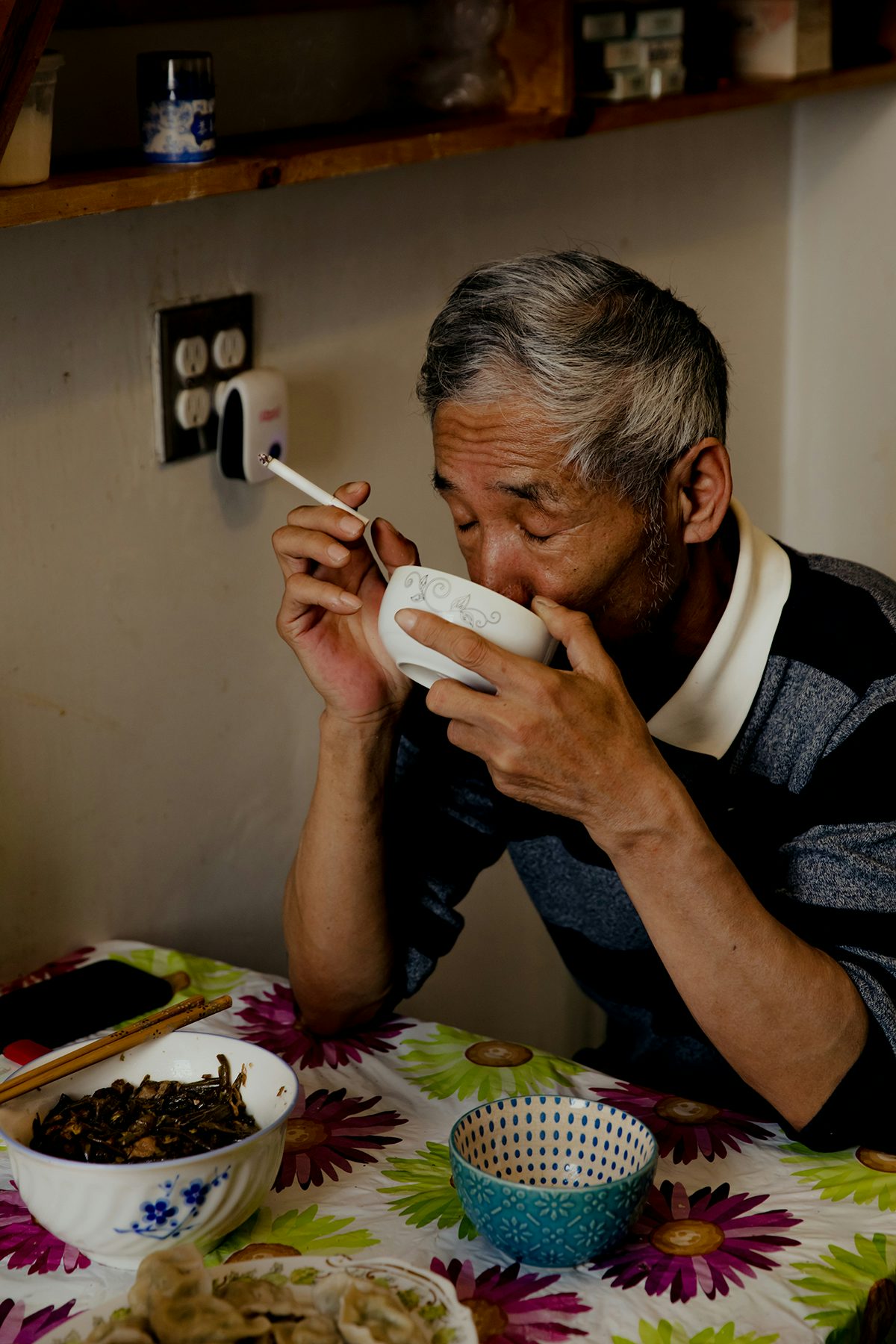 Image by Justin J Wee showing Mr Gao in his kitchen holding a cigarette in one hand and drinking from a bowl held in the other hand