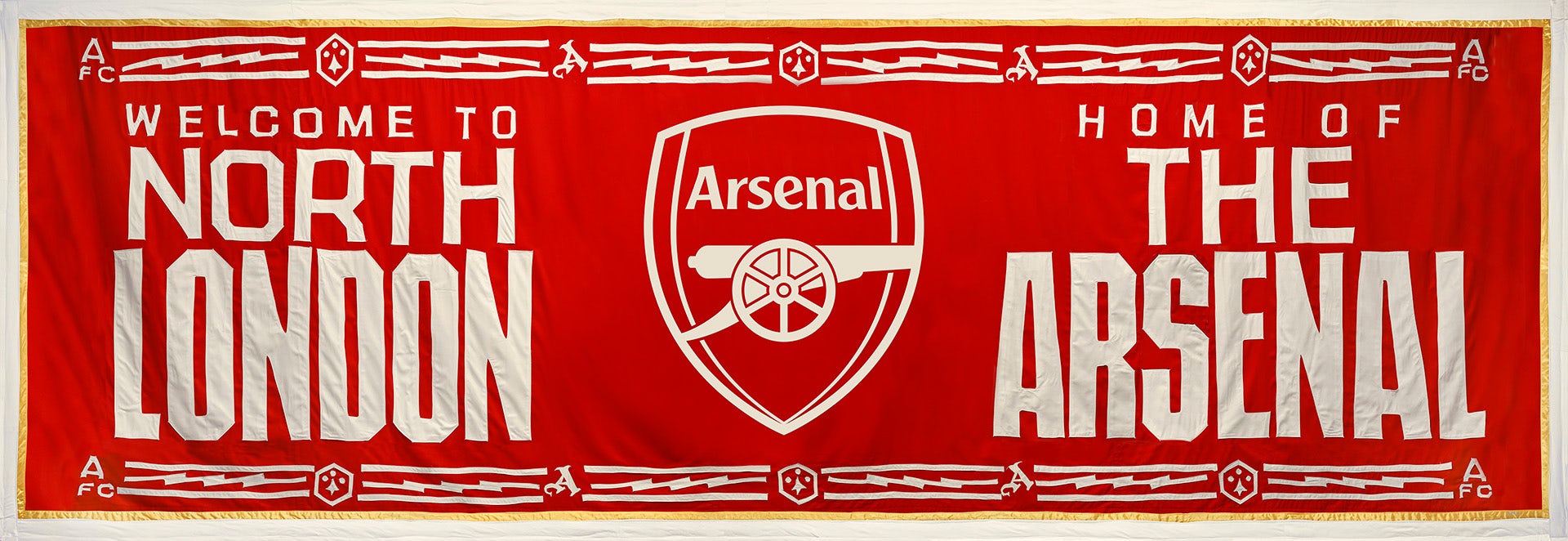 Image shows an Arsenal banner by Jeremy Deller featuring the club's cannon crest in the centre. The banner reads 'Welcome to North London' and 'Home of the Arsenal' on a red background