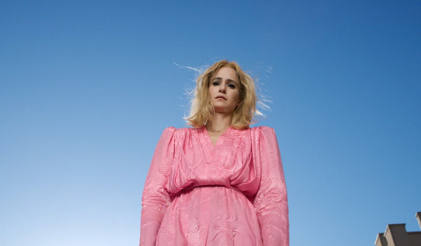 Image by Alex Prager shows a photograph of the character Cecily, who has blonde hair and is wearing a pink dress with a puffy torso and sleeves, stood against a bright blue sky backdrop