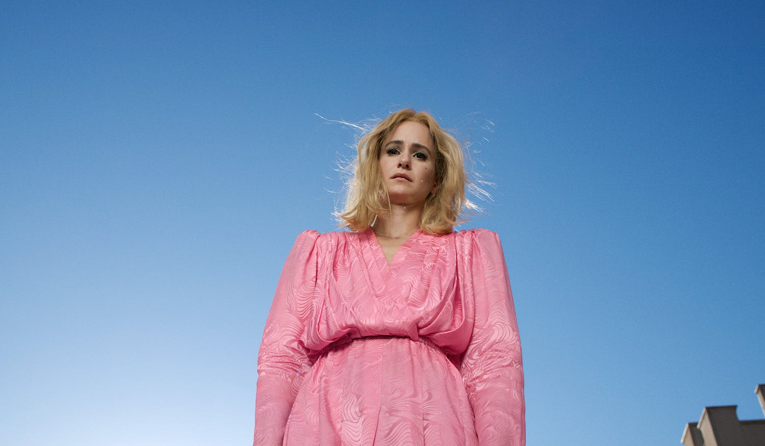 Image by Alex Prager shows a photograph of the character Cecily, who has blonde hair and is wearing a pink dress with a puffy torso and sleeves, stood against a bright blue sky backdrop