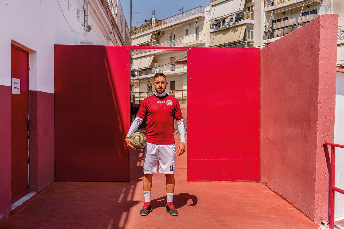 Image by Niko J. Kallianiotis shows a person wearing a red and white football kit holding a football under one arm, stood within an enclosed outdoor space with red walls