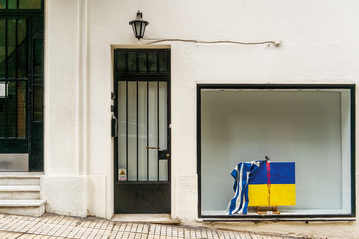 Image by Niko J. Kallianiotis shows a sloping street with a door next to a large window, which shows an object draped in the Greek and Ukrainian flags