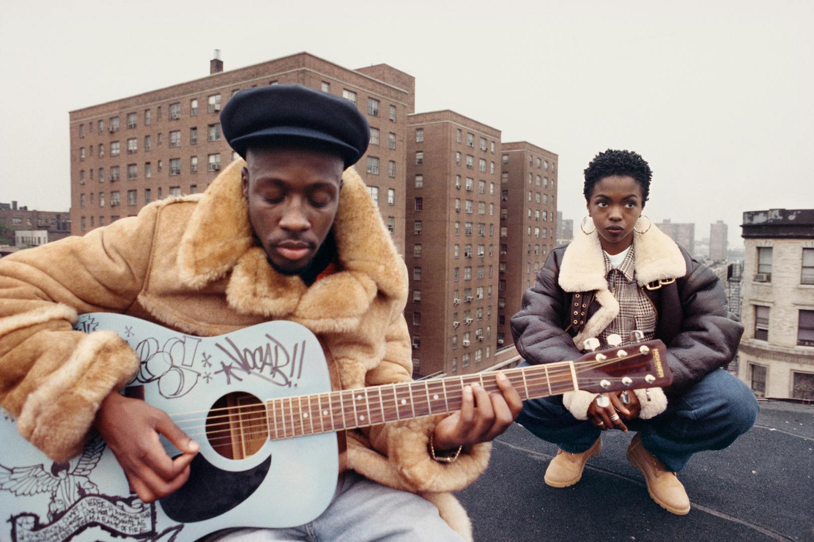 Photograph of Wyclef Jean playing a signed blue acoustic guitar wearing a furry coat and black hat, next to Lauryn Hill wearing an aviator-style jacket. Apartment buildings are in the background