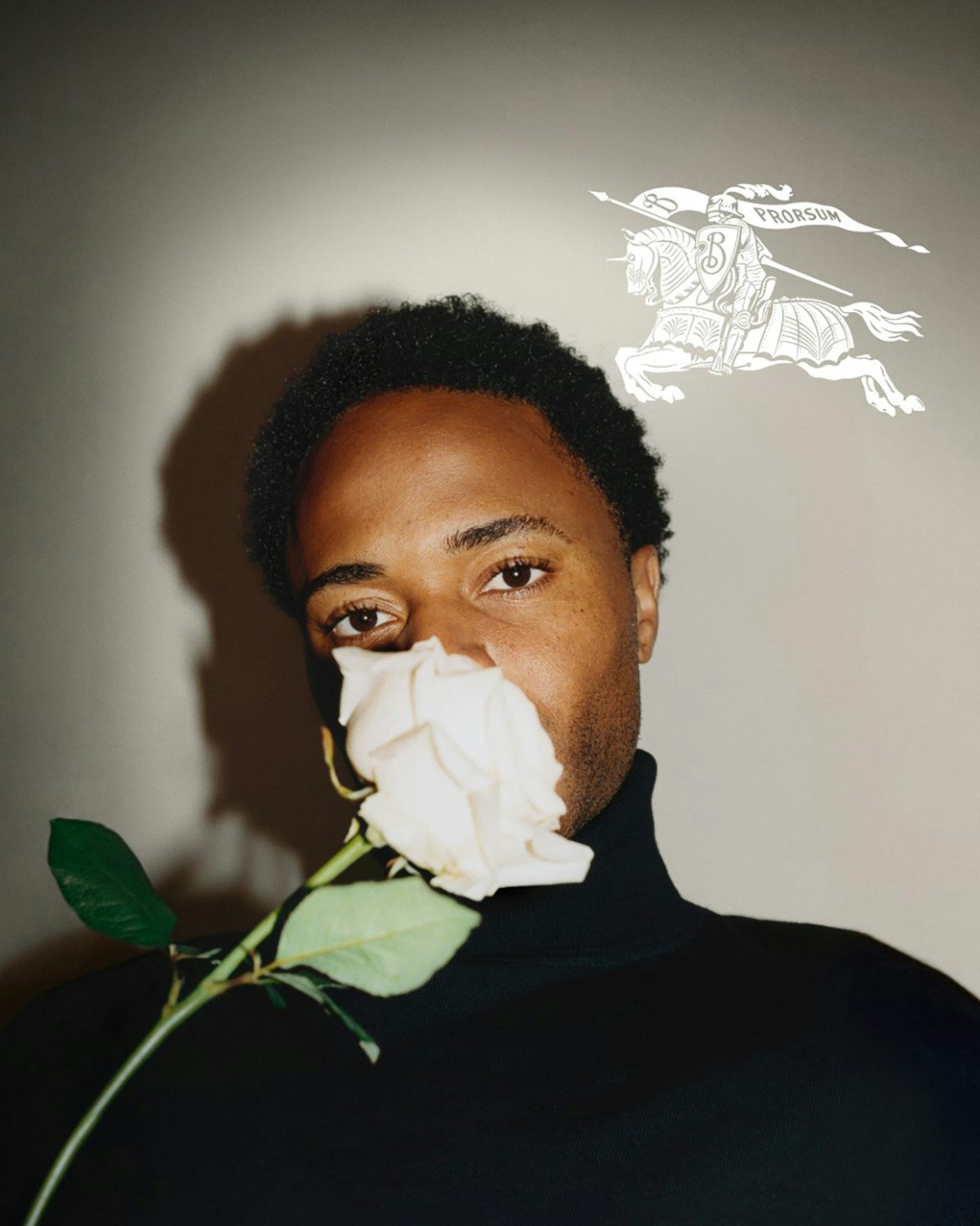 Portrait photo of Raheem Sterling wearing a dark roll neck top holding a white rose to his face. On top of his photo is the new Burberry logo in white, showing a knight riding a horse and carrying a flag that reads 'Prorsum'