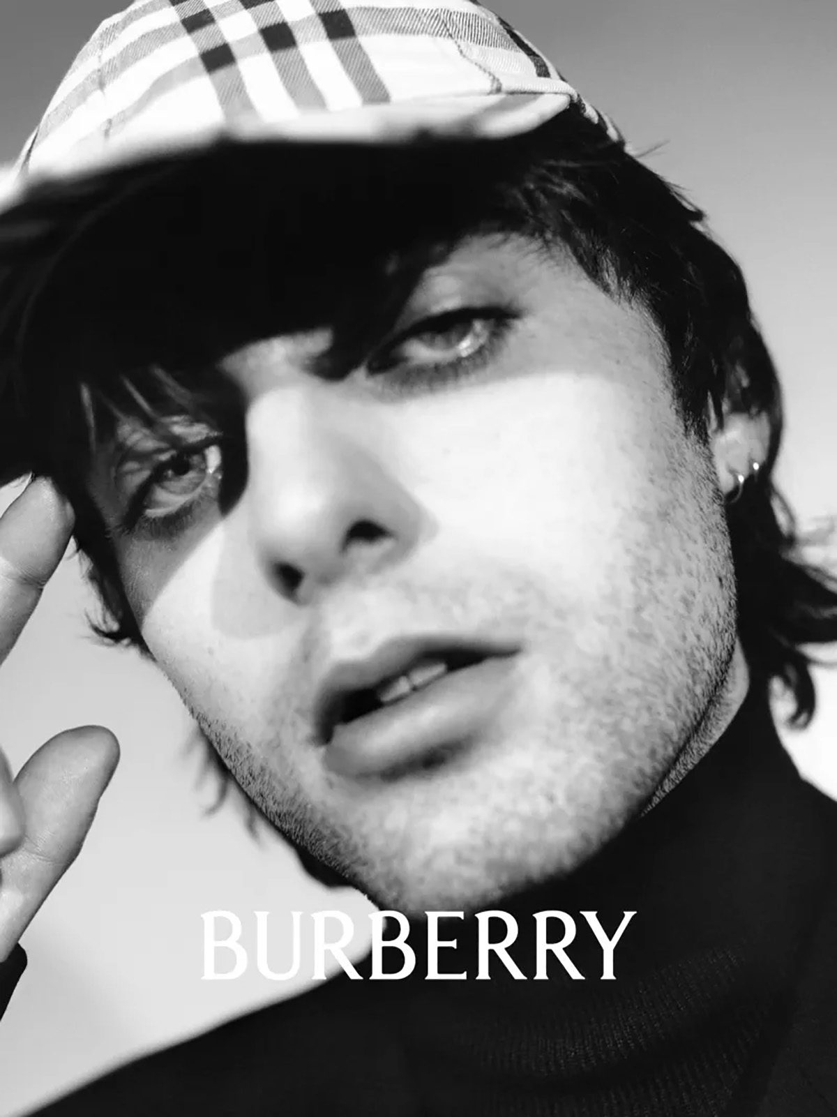 Black and white portrait shot by Tyrone Lebon of Lennon Gallagher wearing a dark roll neck top and a Burberry check cap, next to the new Burberry wordmark