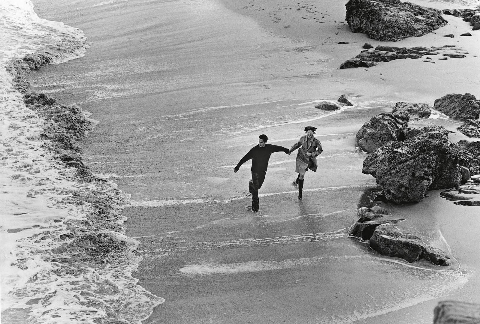 Black and white photograph of Stella Tennant and Andrea Boccaletti running on a beachfront on the set of the Burberry Spring Summer 1999 campaign
