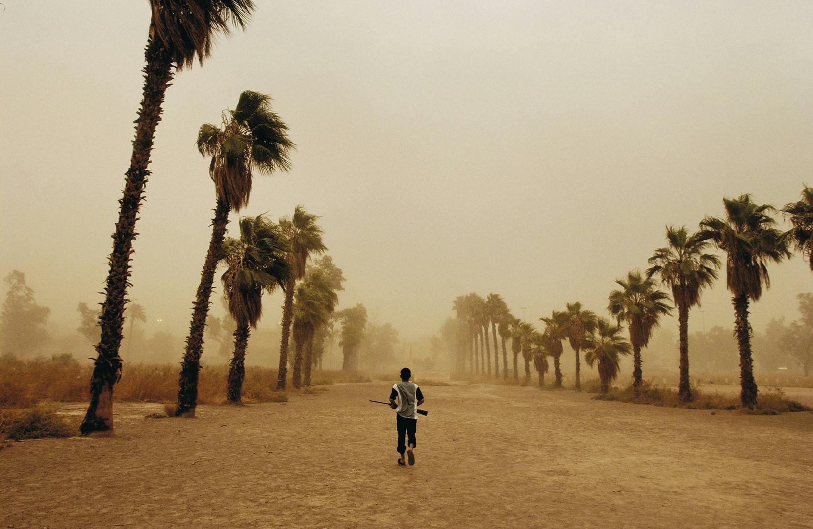 Photograph of a young person holding a gun while walking away from the camera in a dusty tree-lined landscape, taken from Glad Tidings of Benevolence by Moises Saman