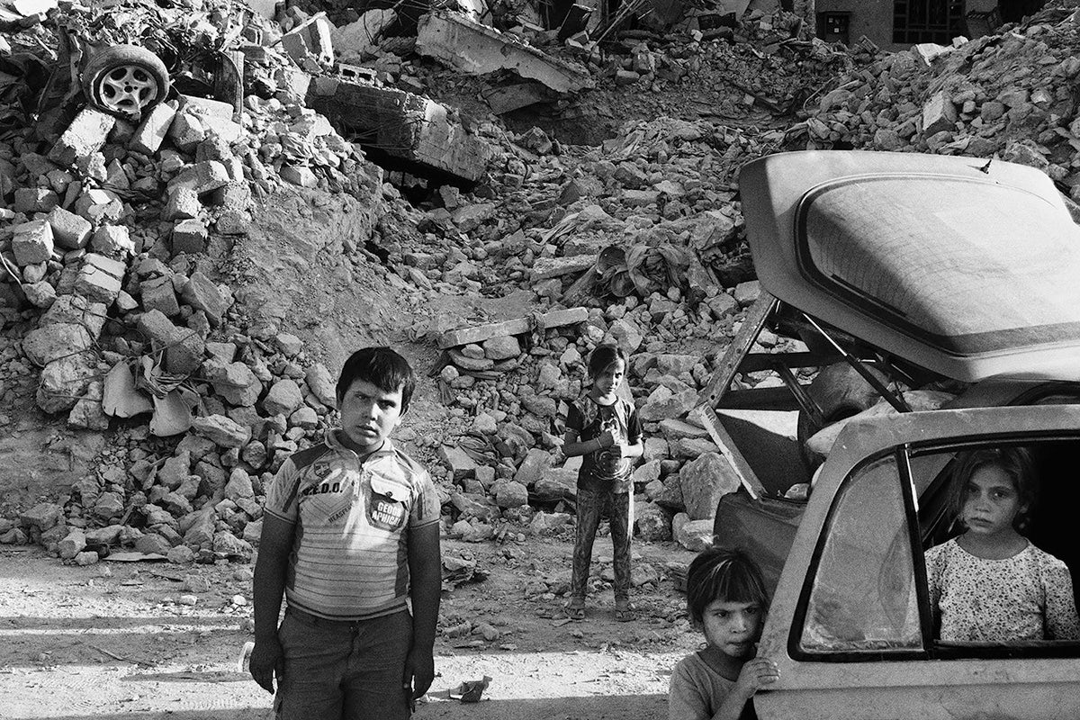 Black and white photograph of four young children in and around a car next to a mound of rubble in Glad Tidings of Benevolence by Moises Saman