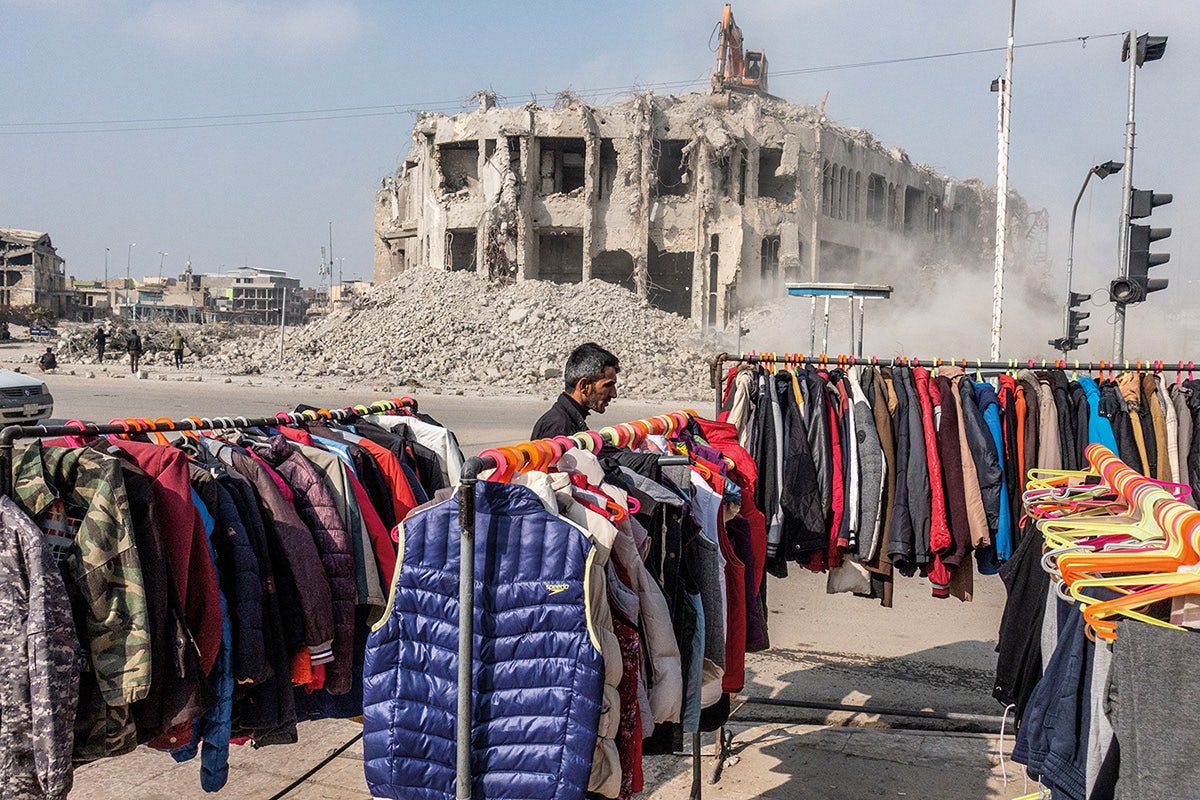 Photograph of a person stood next to rails of clothing with a dilapidated building in the background, featured in Glad Tidings of Benevolence by Moises Saman