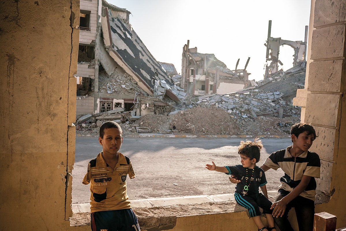 Photograph of three children, including one child who appears to have no arms, sat in the entry way to a building with dilapidated building in the background, taken from Glad Tidings of Benevolence by Moises Saman
