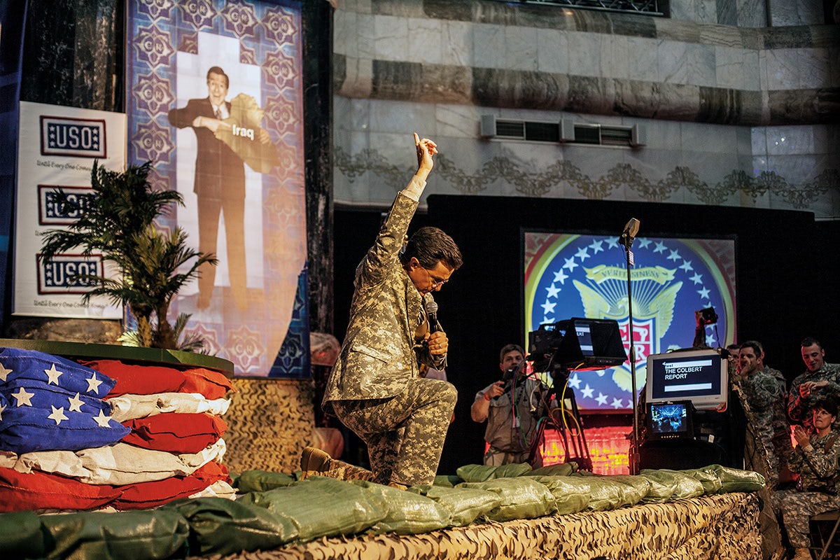 Photograph of a person wearing a military outfit kneeling on one knee with a hand raised in the air and the other holding a microphone, taken from Glad Tidings of Benevolence by Moises Saman