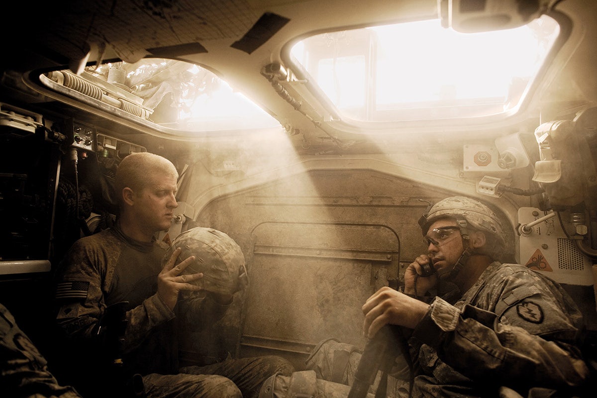 Photograph of two soldiers sat inside a sun-filled army tank. One is examining a helmet, the other is on the phone, taken from Glad Tidings of Benevolence by Moises Saman