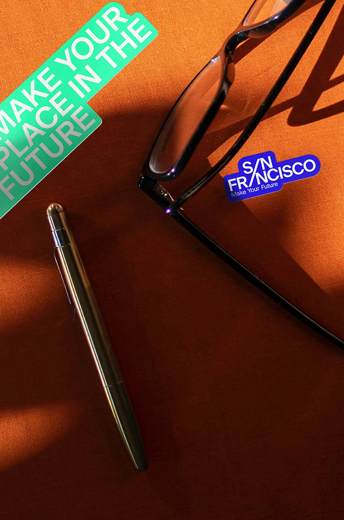 Photograph showing the San Francisco place branding on an orange table with stickers that read 'Make your place in the future' and 'San Francisco' next to a pair of glasses and a pen