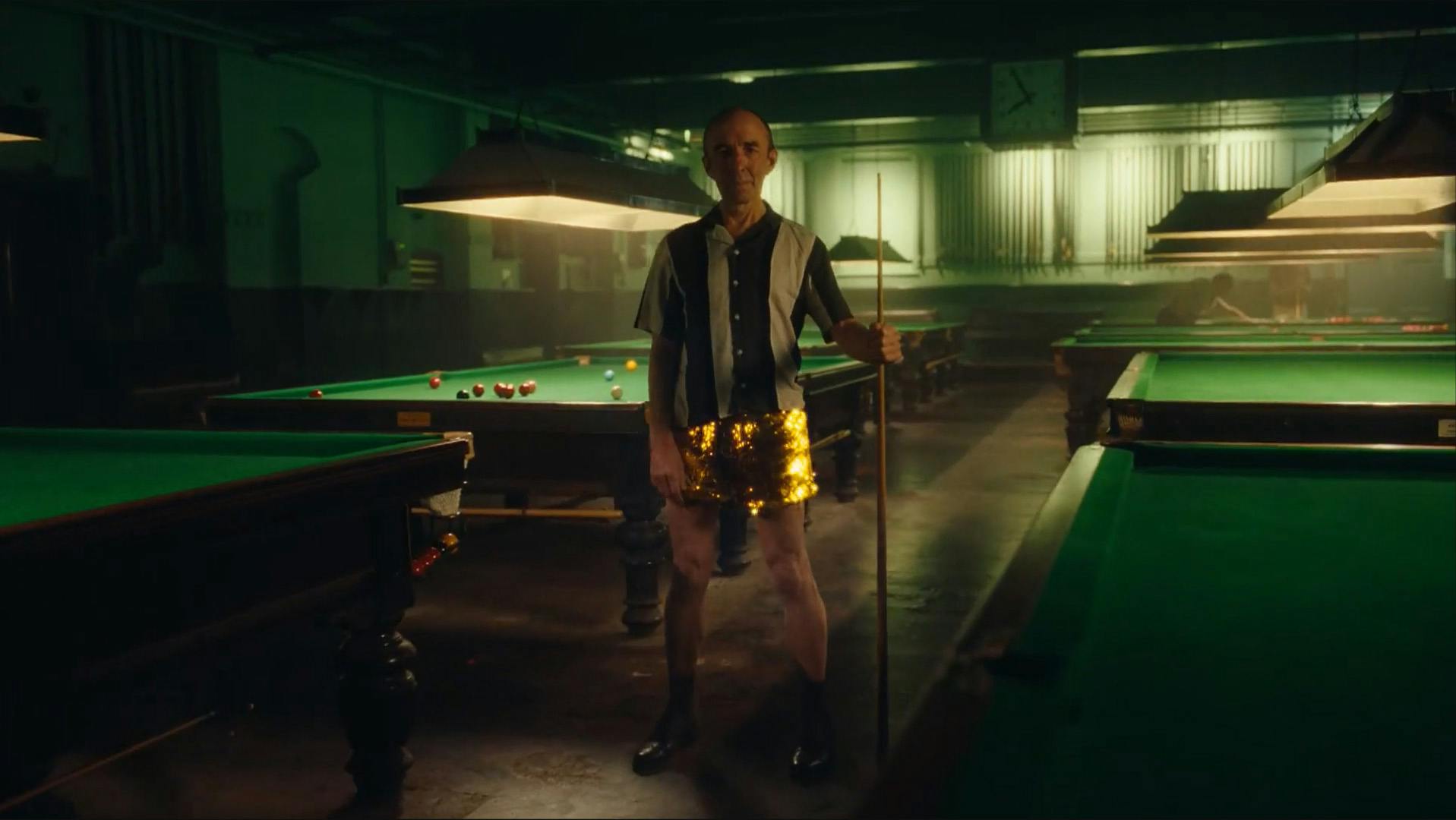 Still from the BBC Eurovision 2023 ad showing a person wearing a button-up shirt and gold glittery shorts holding a pool cue in a darkened empty pool bar