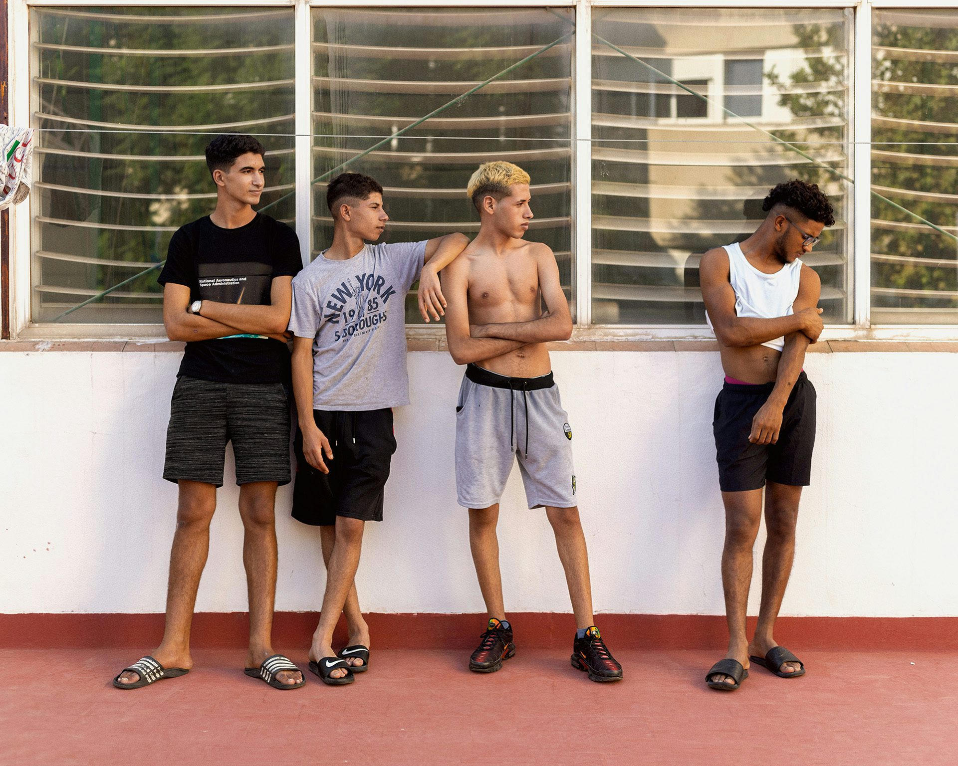 Photograph taken from Dialect by Felipe Romero Beltran of four young people stood outside in front of windows, in an area with a red floor. The three people on the left are stood close to one another, while the fourth on the right, wearing a white crop top and dark shorts, is stood apart
