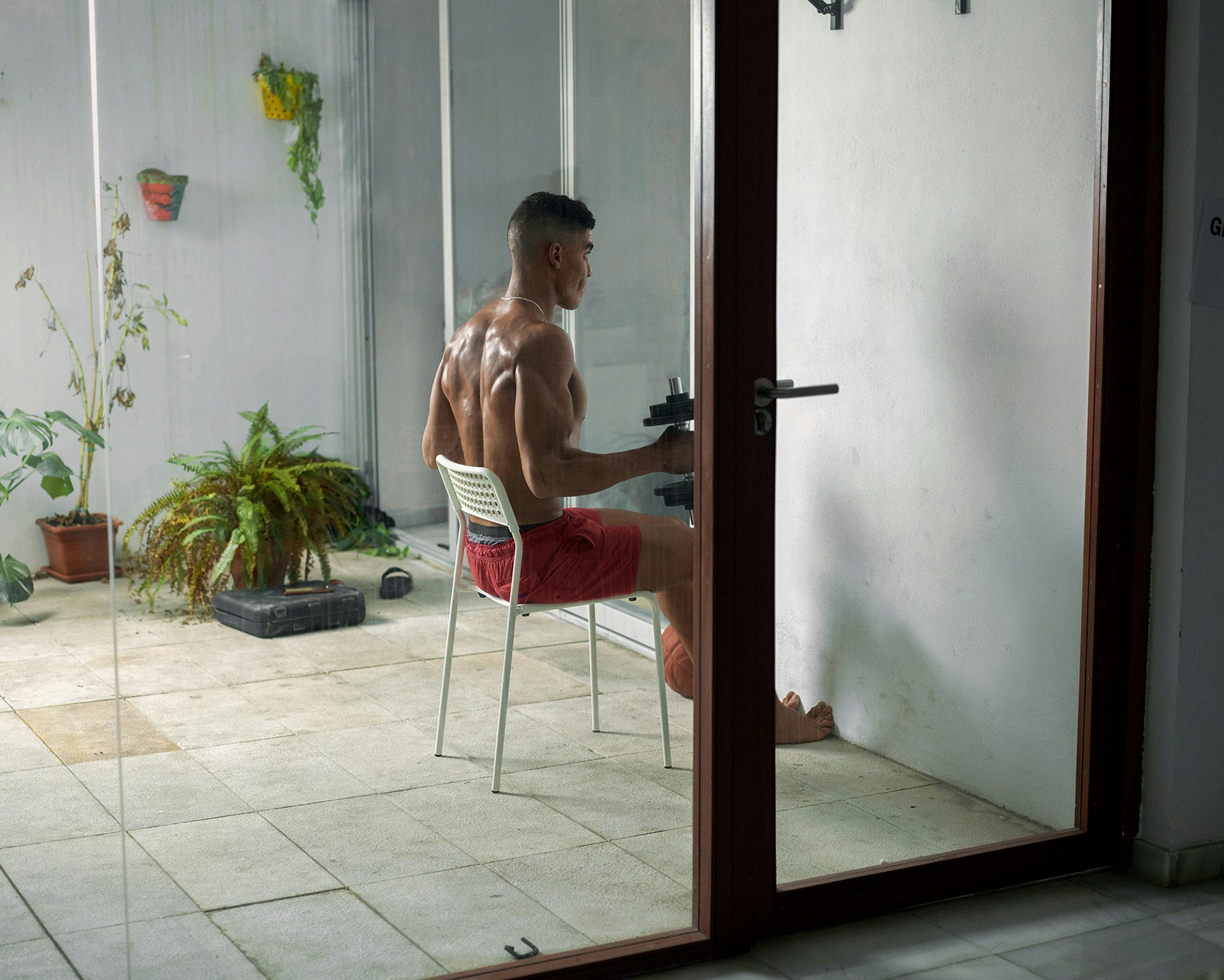 Photograph from Dialect by Felipe Romero Beltran showing a young person, who is wearing red shorts, holding arm weights while sat down on a garden chair, as soon through a glass window and door