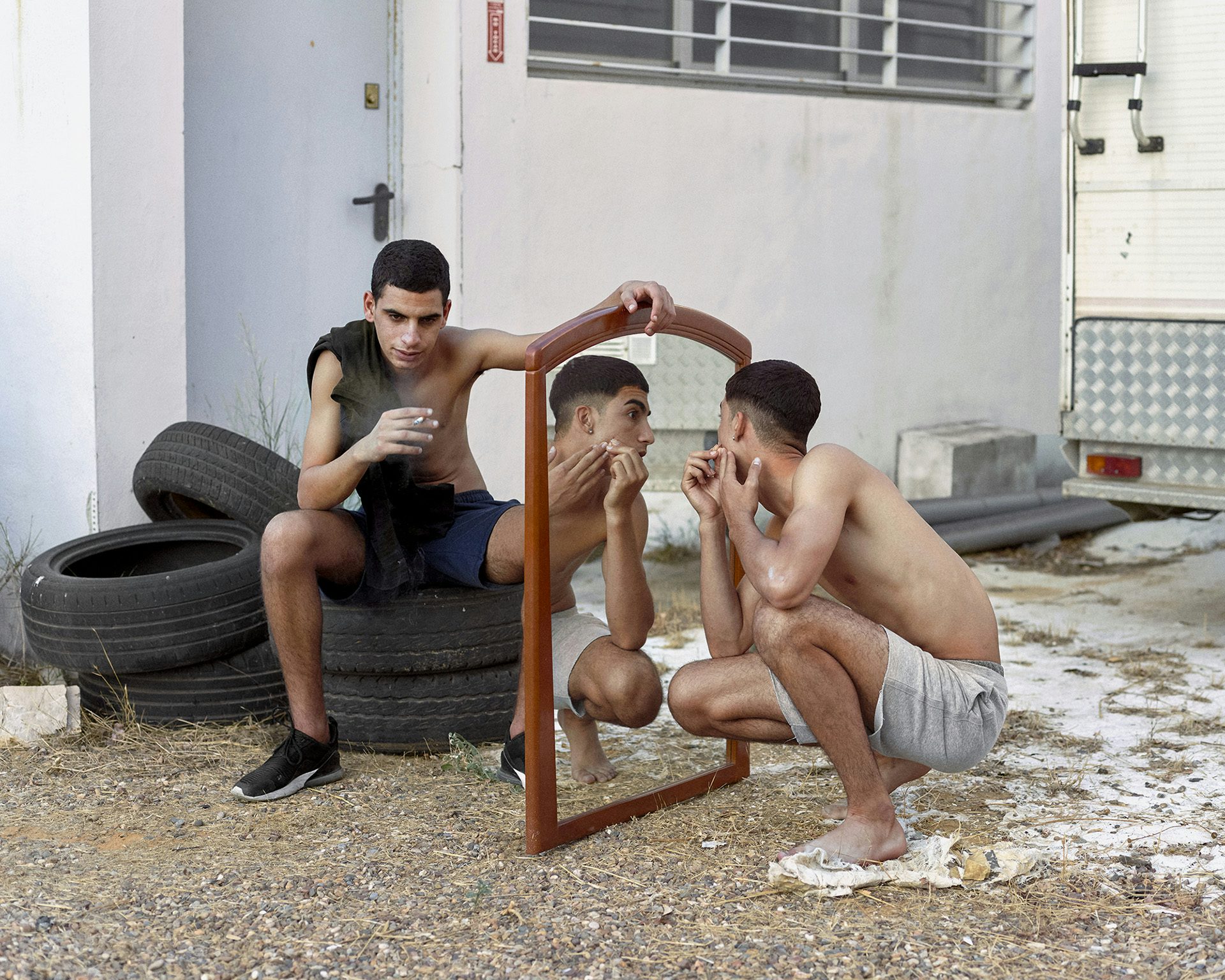 Photograph of a young person sat on a pile of car tyres smoking a cigarette and looking at the camera, holding a mirror that another young person wearing grey shorts is looking into, their reflection visible. The image is taken from Dialect by Felipe Romero Beltran