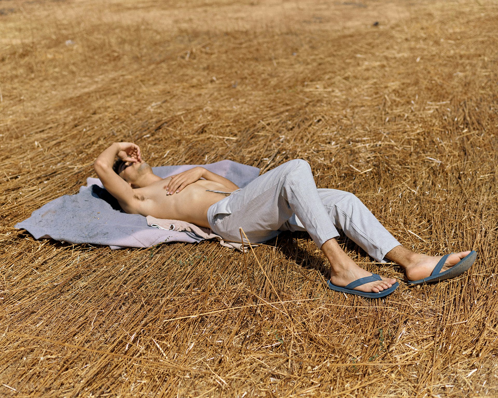 Photograph of a person wearing grey tracksuit bottoms and flip flops lying down on a brown dry surface, holding their arm to shield their face, taken from Dialect by Felipe Romero Beltran