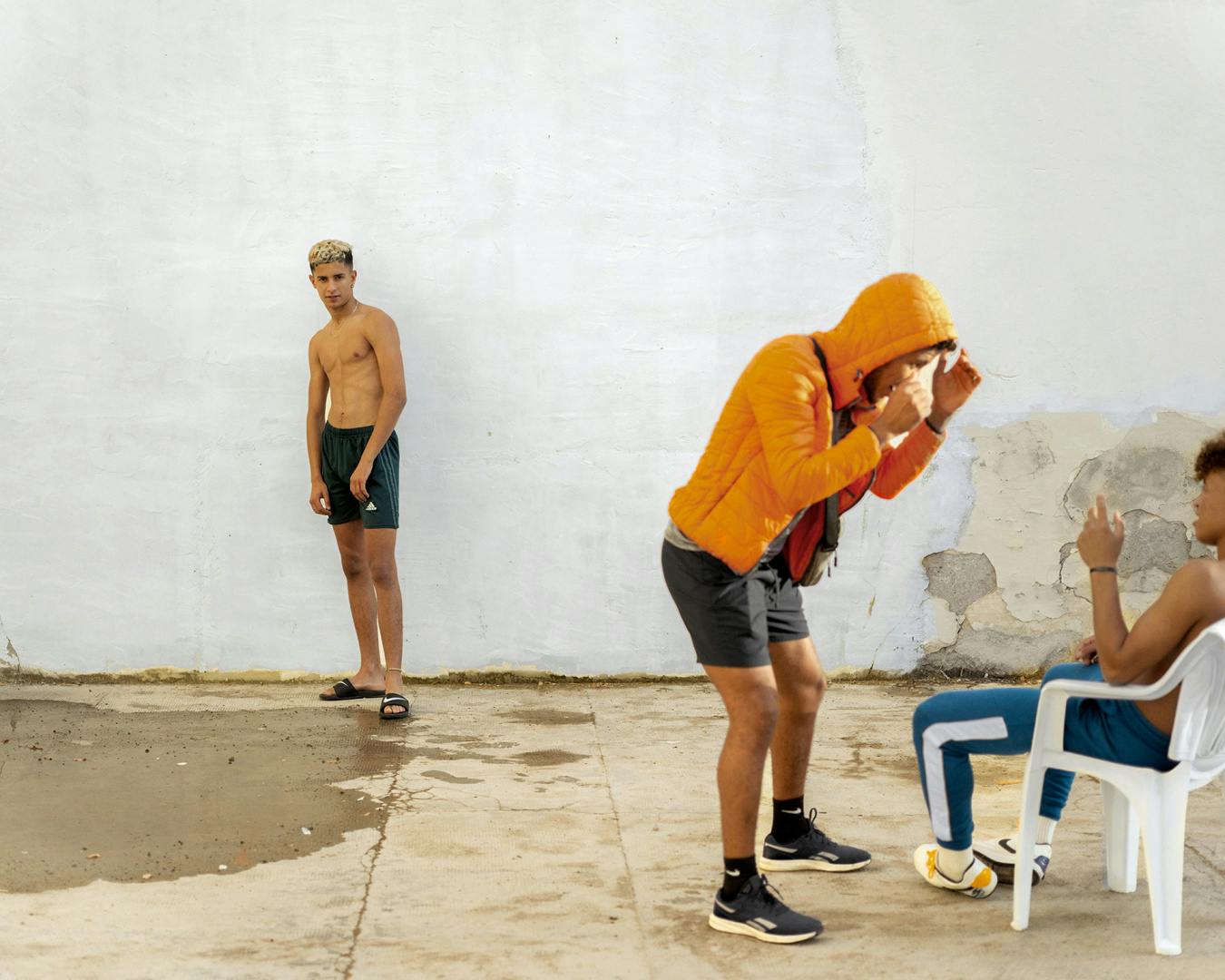Photograph of a young person wearing an orange hooded jacket and dark shorts holding their hands up to their face and leaning over another young person wearing blue sportswear sat down in a white plastic chair. A third person wearing black shorts and pool sliders is stood against a white wall watching on, taken from Dialect by Felipe Romero Beltran