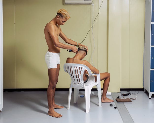 Photograph of a young person wearing white underwear and no shirt, who is cutting the hair of another person sat in a white plastic chair with their back to the camera, taken from Dialect by Felipe Romero Beltran