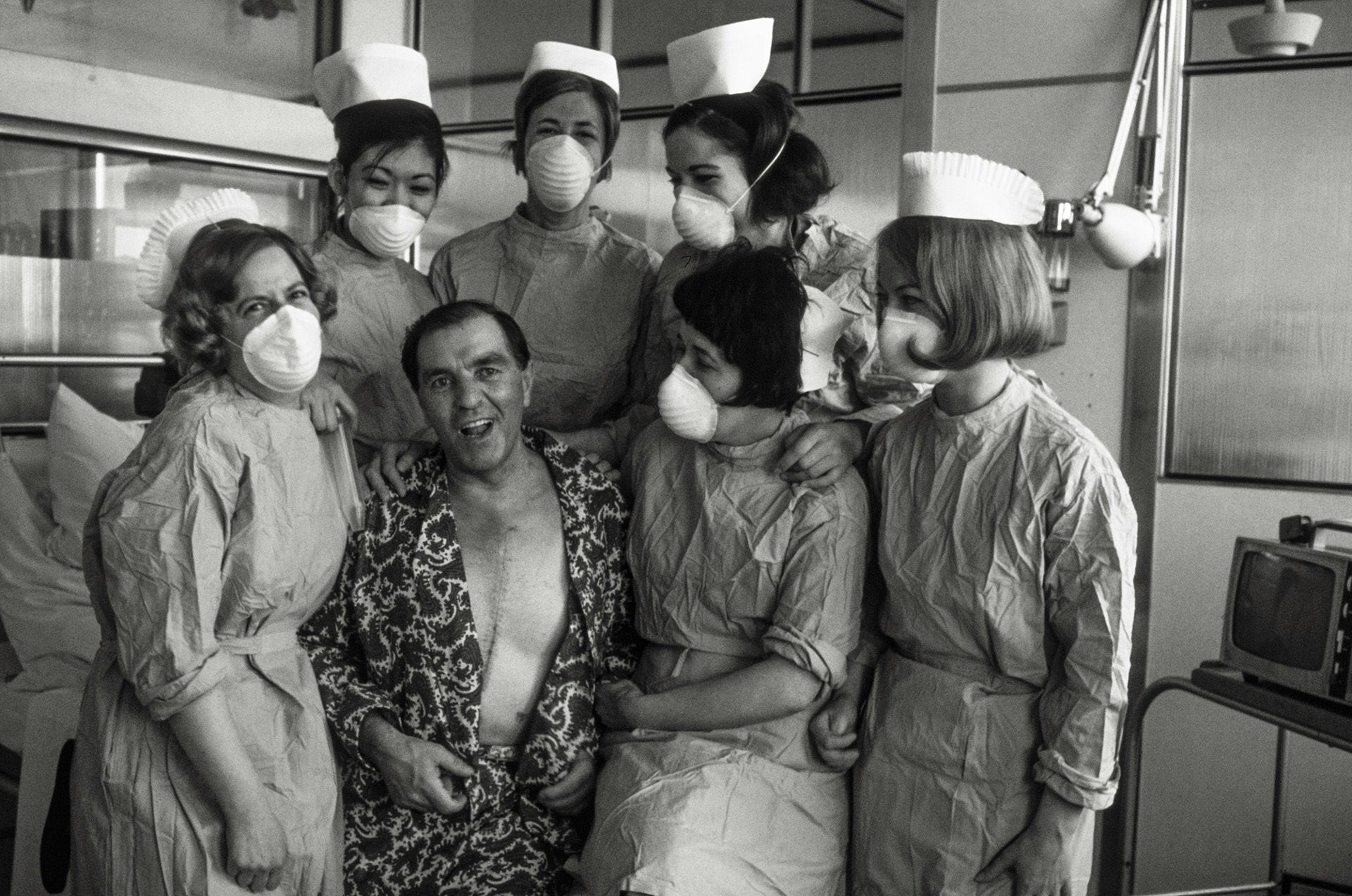 Black and white photograph of a group of NHS nurses smiling with a masked patient who is wearing a patterned robe