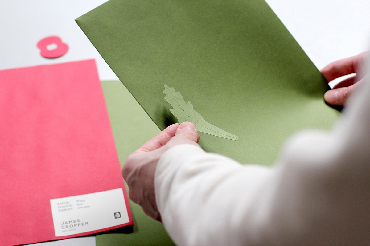 Image shows a person holding a leaf cut-out against green paper next to red paper as part of the redesign of the Royal British Legion poppy