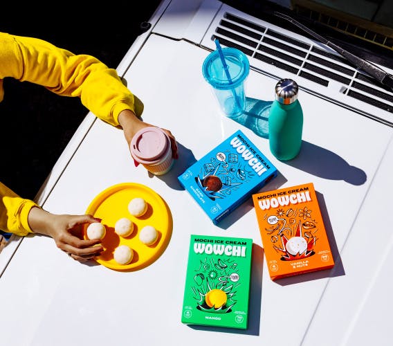 Image shows a table topped with a plate of mochi balls and drinkware, next to blue, orange and green boxes of Wowchi mochi with line illustrations and mochi balls on the front