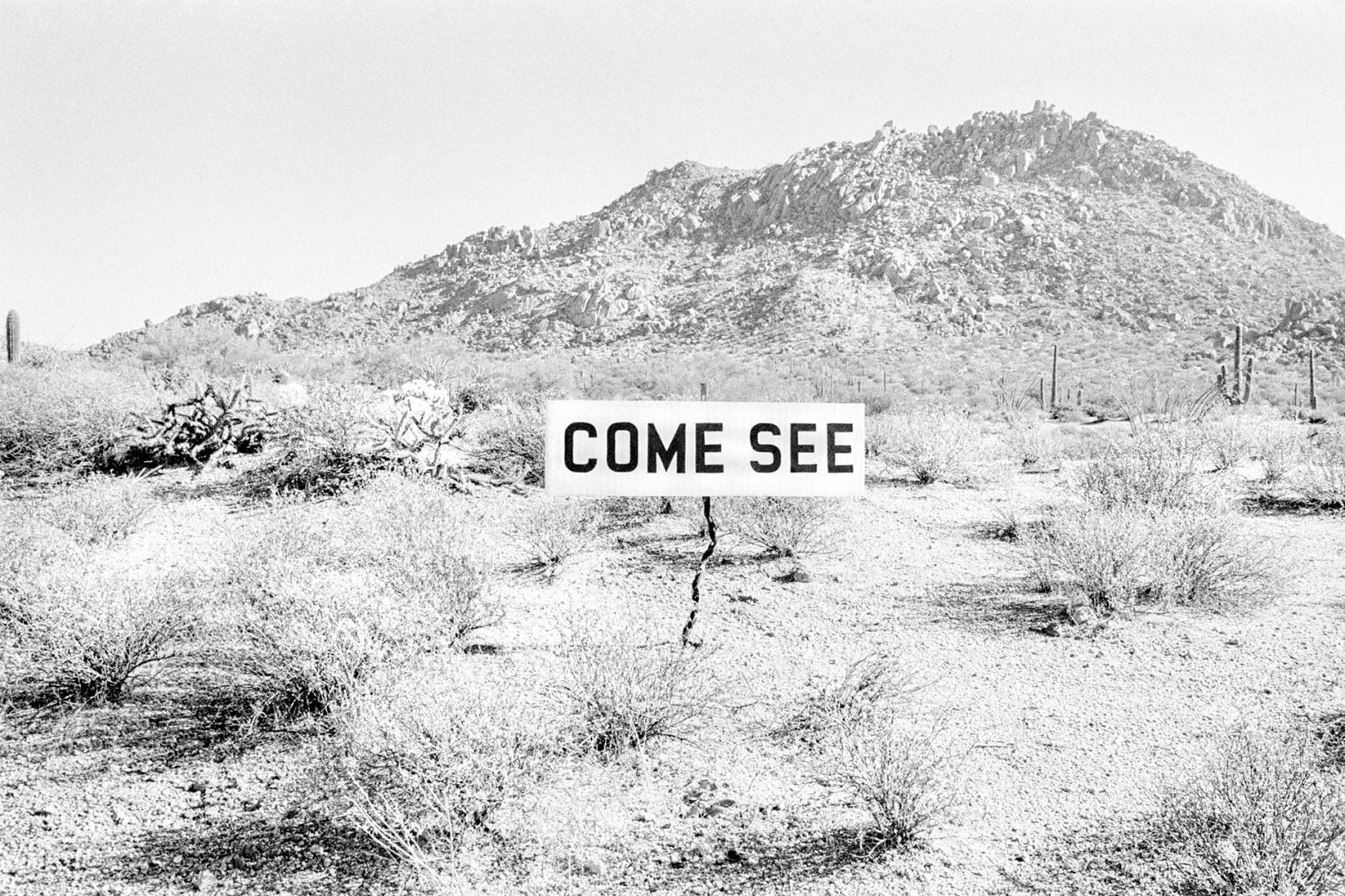 Black and white photograph of a signpost in the desert that reads 'Come see', taken from the book 'David Hurn Photographs 1955-2022'