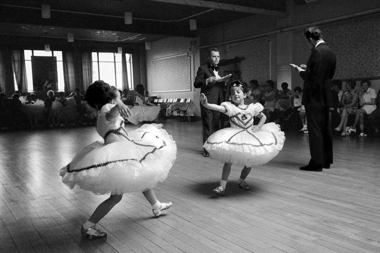 Black and white photograph of two children wearing frilly dresses dancing in a ballroom dance competition, with two people wearing suits and holding clipboards behind them, taken from the book 'David Hurn Photographs 1955-2022'