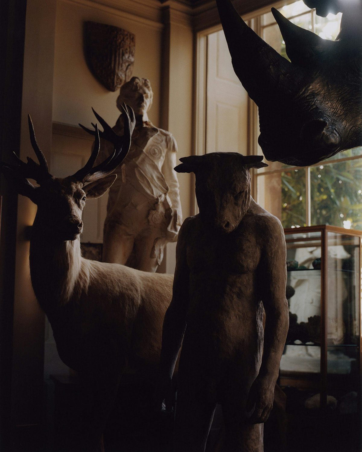 Photograph taken from Folly by Jamie Murray showing a taxidermy deer, rhinoceros head, and a human-like figure with an animal's head, inside a darkened neutral room