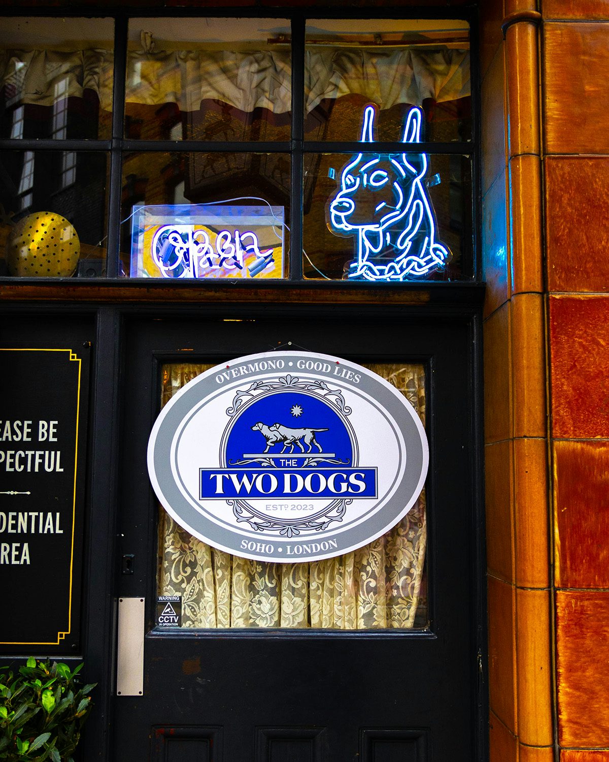 Image shows the an oval poster featuring two illustrated Dobermanns and labelled The Two Dogs, as well as neon lighting in the shape of a dog's head, in the window of Overmono's pop up pub