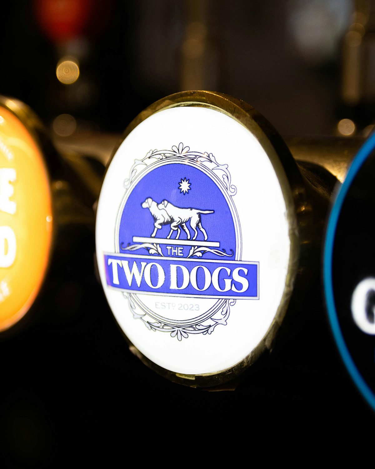 Image shows a pub keg badge for Overmono's pop-up beer brand Two Dogs, featuring a blue background and an illustration of two dogs
