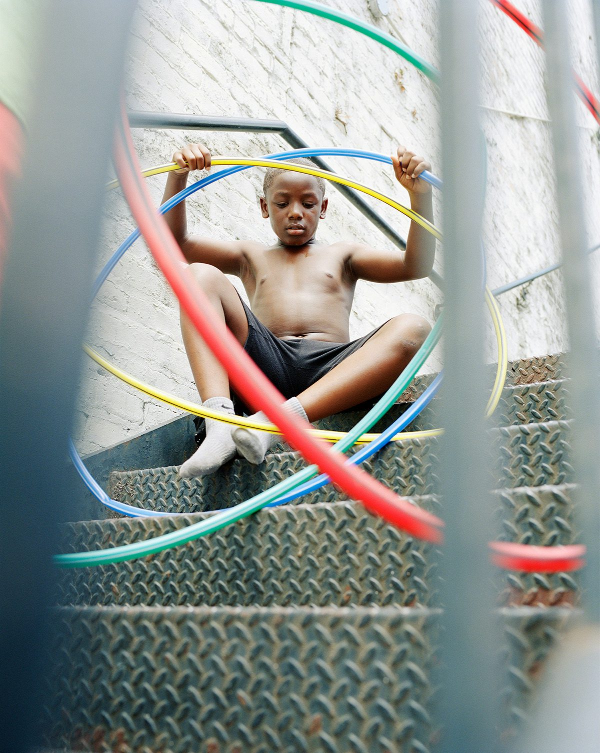 Photograph by Vasantha Yogananthan of a young person wearing shorts and socks, sat on a metal staircase with colourful hula hoops surrounding them. The scene is shot through the metal bars of the staircase which are visible in the frame